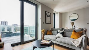 A modern living room with a grey sofa, a coffee table, and a view of a cityscape through large windows. - Student Only Clay Wembley (Wembley)