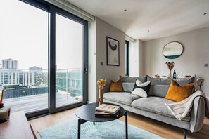 Ein modernes Wohnzimmer mit grauem Sofa, einem Couchtisch und Blick auf eine Stadtlandschaft durch große Fenster.
