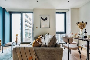A modern living room with a sofa, a dining table set for two, and a large window with a view of a building. - Student Only Clay Wembley (Wembley)