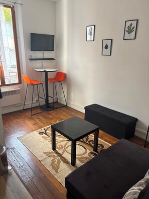 Ein kleines, möbliertes Zimmer mit einem Bett, einem Tisch, zwei Stühlen, einem Fernseher und gerahmten Bildern an der Wand.