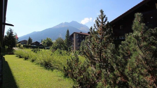 Un bâtiment avec une montagne en arrière-plan, entouré de verdure et d’arbres.