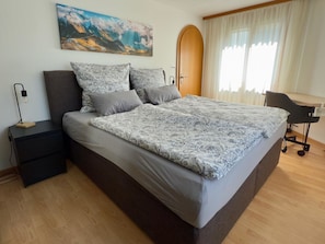Ein modernes Schlafzimmer mit einem großen Bett, einem Nachttisch, einem Schreibtisch und einem Fenster mit Vorhängen.