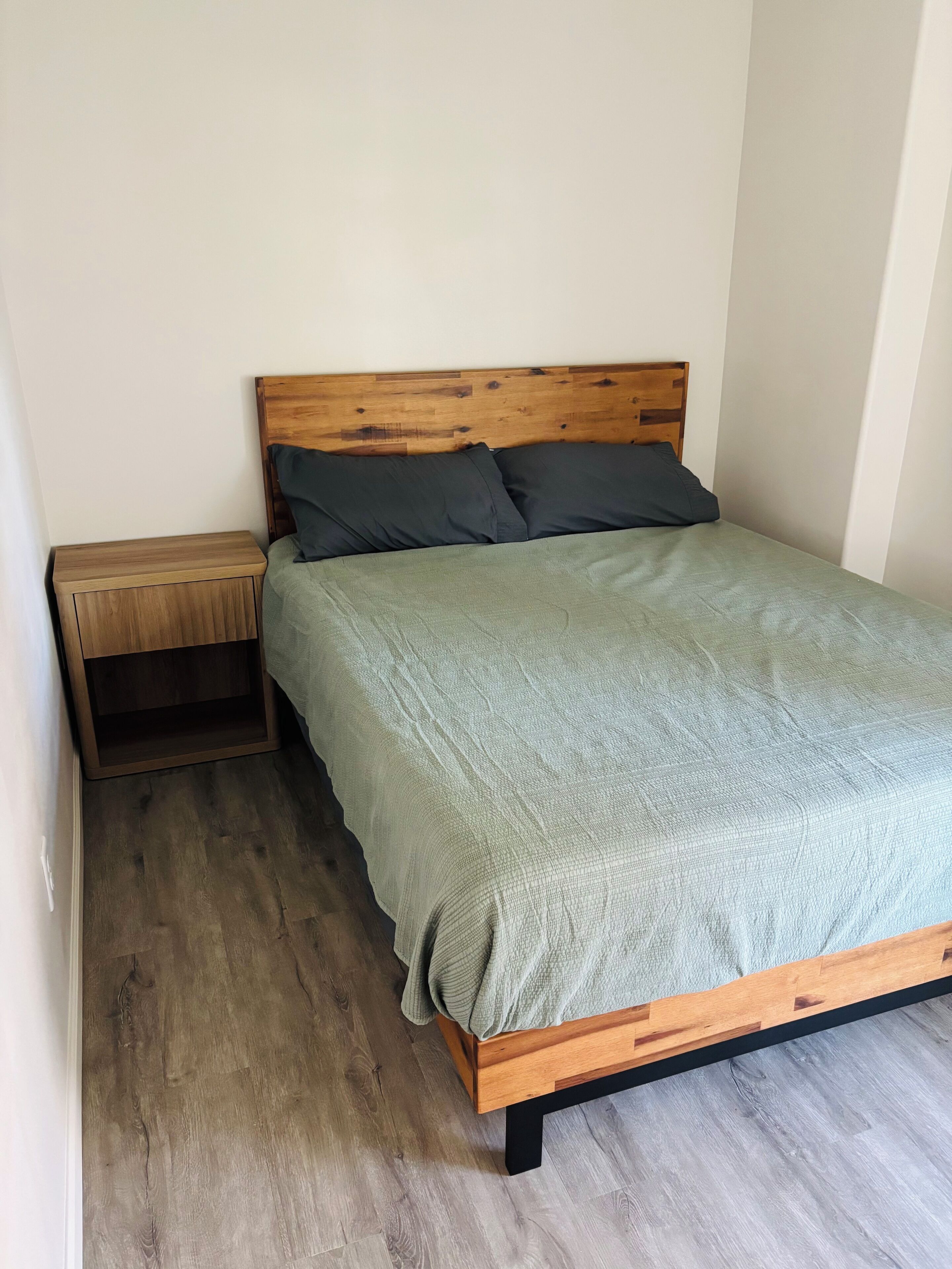 Un lit simple avec une tête de lit en bois et des oreillers foncés, une table de chevet avec un tiroir et un plancher en bois de couleur claire.