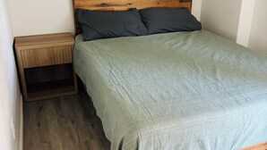 Un lit simple avec une tête de lit en bois et des oreillers foncés, une table de chevet avec un tiroir et un plancher en bois de couleur claire.