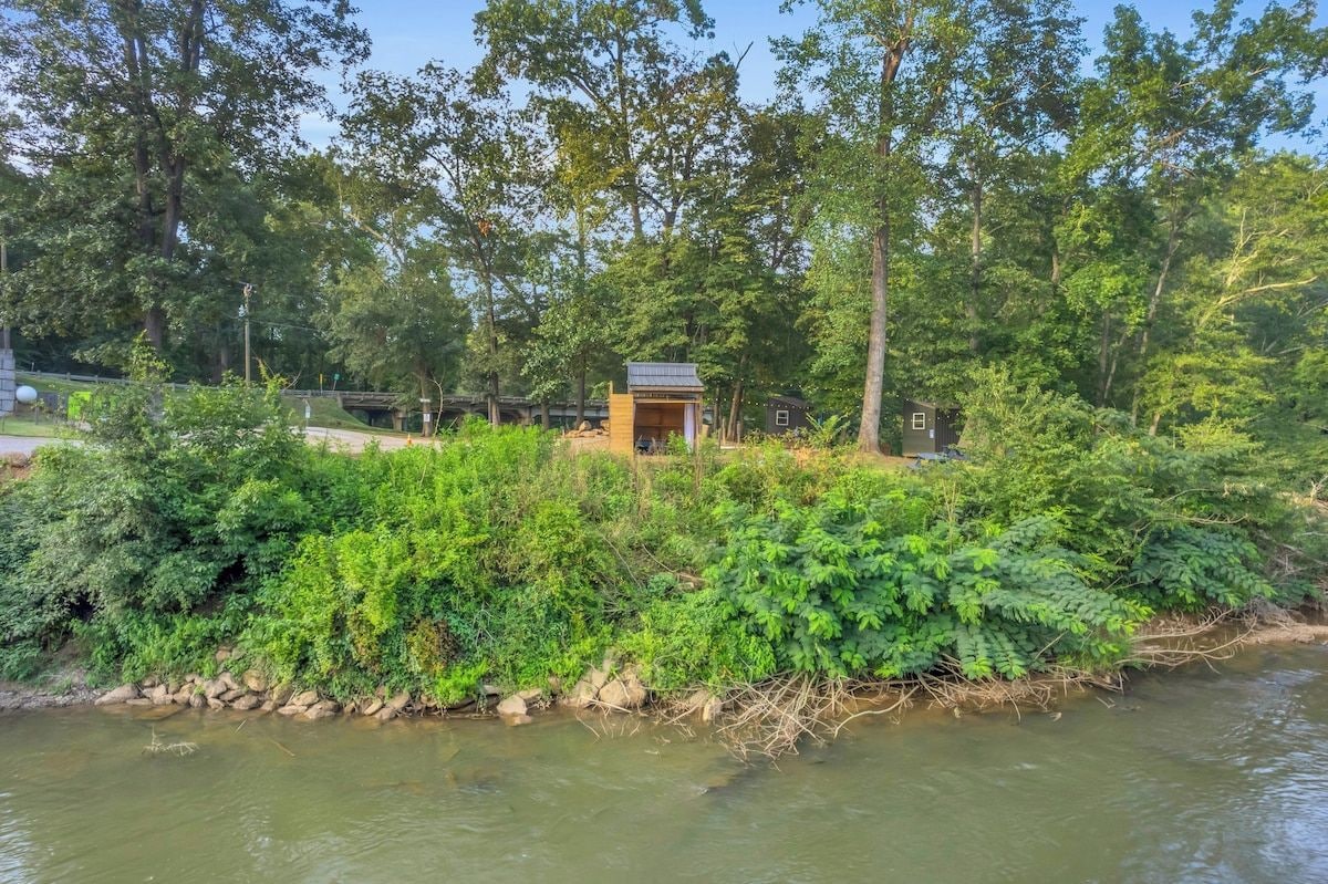En lille hytteagtig bygning omgivet af frodig vegetation og et vandareal.