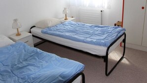 Een eenvoudige slaapkamer met twee eenpersoonsbedden, een nachtkastje, een lamp en een radiator.