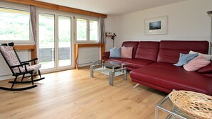 Ein Wohnzimmer mit einem roten Sofa, einem Glastisch, einem Schaukelstuhl und einem Korbstuhl.