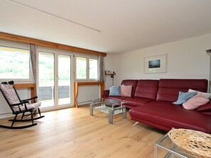Ein Wohnzimmer mit einem roten Sofa, einem Glastisch, einem Schaukelstuhl und einem Korbstuhl.