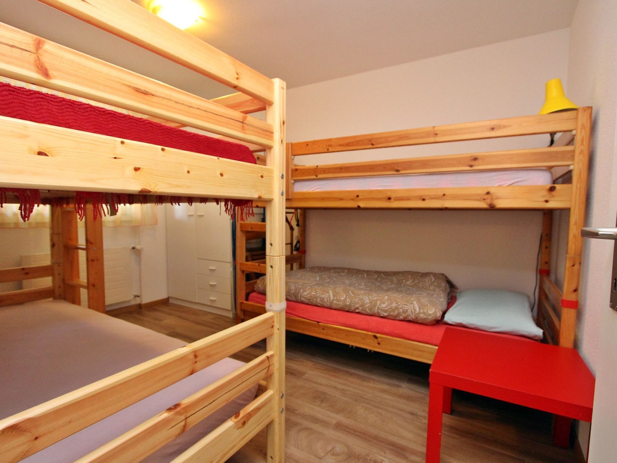 A bunk bed room with a wooden bunk bed, a red table, and a white wardrobe.