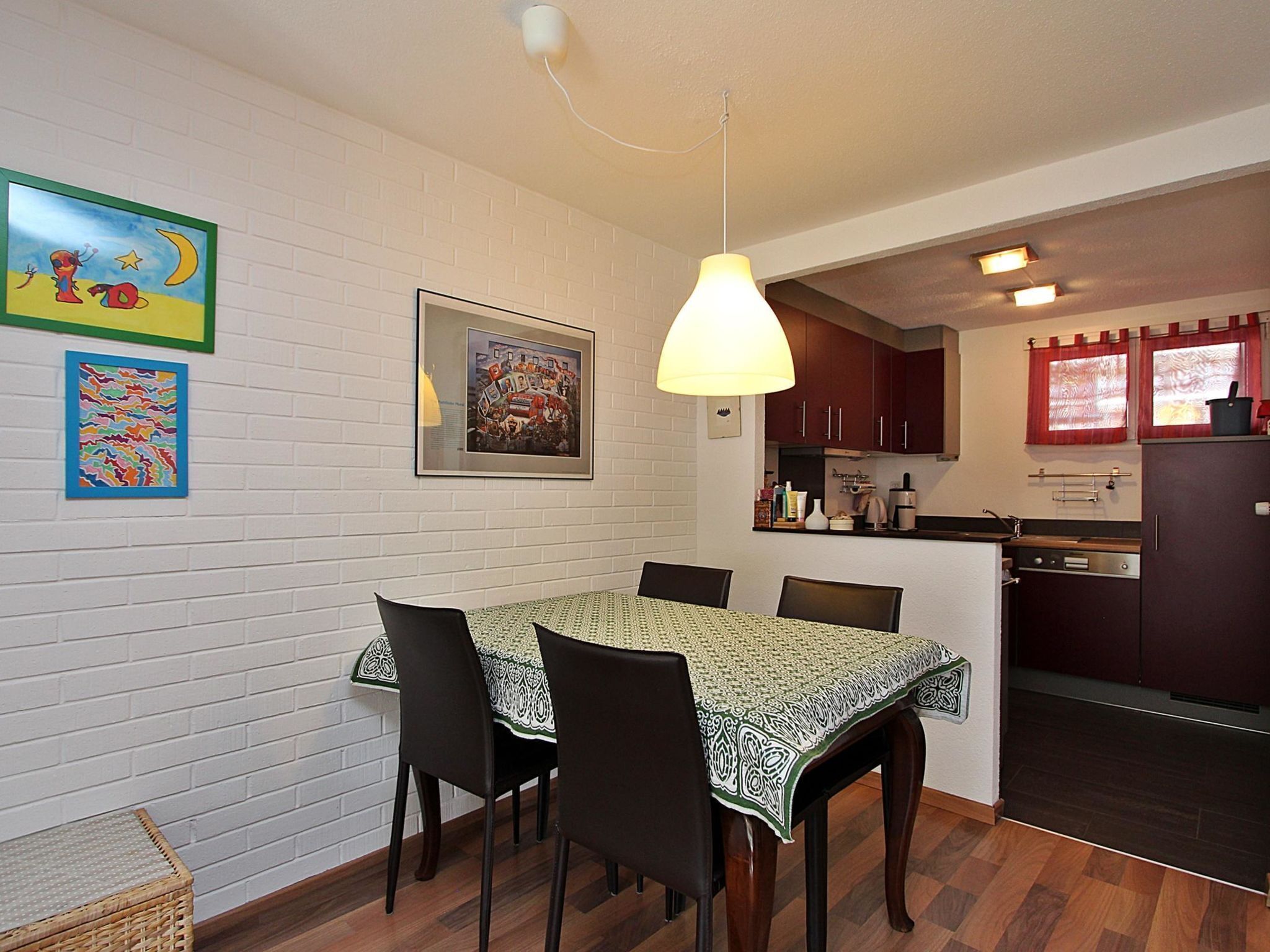 A dining area with a table and chairs, a hanging light fixture, and a kitchen in the background.