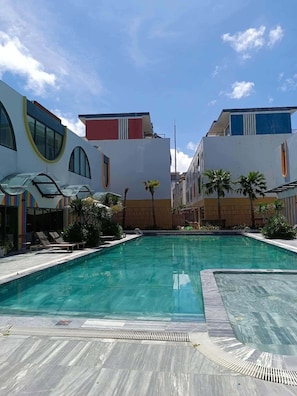 Una piscina con solárium de baldosas, rodeada de edificios con grandes ventanales y un cielo despejado.