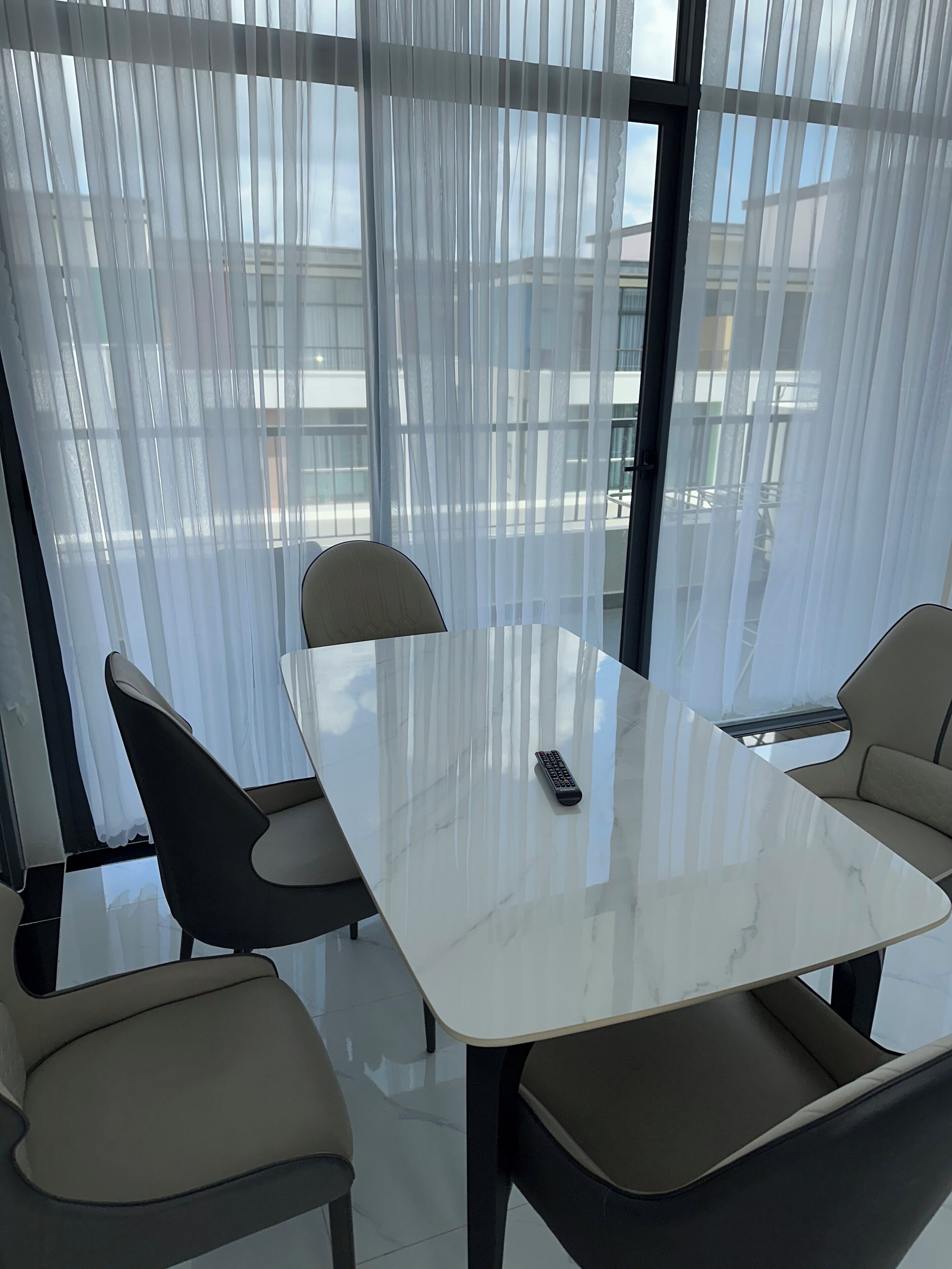 Ein moderner Konferenzraum mit Glastisch und vier Stühlen, großen Fenstern mit durchsichtigen Vorhängen und Blick auf ein Gebäude.
