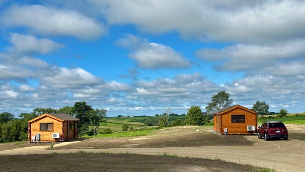 Un entorno rural con dos cabañas de madera, una con garaje adjunto, y un coche rojo estacionado cerca.