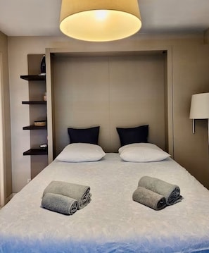 A hotel room with a bed, two pillows, a bedside table, a lamp, and a shelf unit. - POINTE CROISETTE TERRACE STUDIO A0B214 (Cannes)