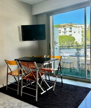 A modern room with a dining table, chairs, a flat-screen TV mounted on the wall, and a balcony with a view of buildings and greenery. - POINTE CROISETTE TERRACE STUDIO A0B214 (Cannes)