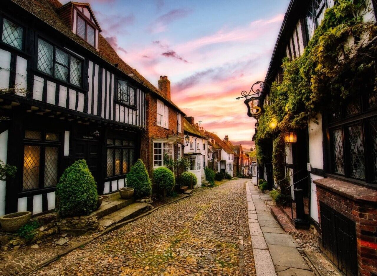 A narrow cobblestone street lined with traditional half-timbered houses, a clear sky, and a sunset.