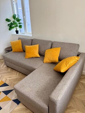 A grey sectional sofa with four yellow cushions, a potted plant, and a geometric patterned rug in a room with a window and wooden flooring. - The Nook in Rye Town Centre (East Sussex)