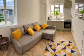 A modern living room with a grey sectional sofa, yellow cushions, a herringbone wood floor, and a geometric patterned rug. - The Nook in Rye Town Centre (East Sussex)