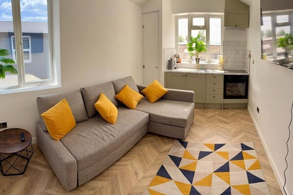 A modern living room with a grey sectional sofa, yellow cushions, a herringbone wood floor, and a geometric patterned rug. - The Nook in Rye Town Centre (East Sussex)