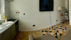 A modern kitchen with a built-in stove, a dining area with a patterned rug, and a flat-screen TV mounted on the wall. - The Nook in Rye Town Centre (East Sussex)