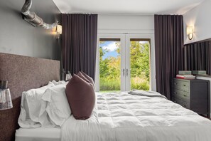 A hotel room with a bed, a bedside lamp, a dresser, and a view of greenery through the door. - Zen Den • Sleeps 14 • 40-Acre Berkshire Retreat (Stephentown)