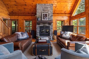 A log cabin interior with a stone fireplace, leather sofas, and a wooden staircase. - Zen Den • Sleeps 14 • 40-Acre Berkshire Retreat (Stephentown)