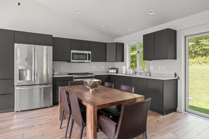 A modern kitchen with stainless steel appliances, a dining table with chairs, and a view of greenery outside. - Zen Den • Sleeps 14 • 40-Acre Berkshire Retreat (Stephentown)
