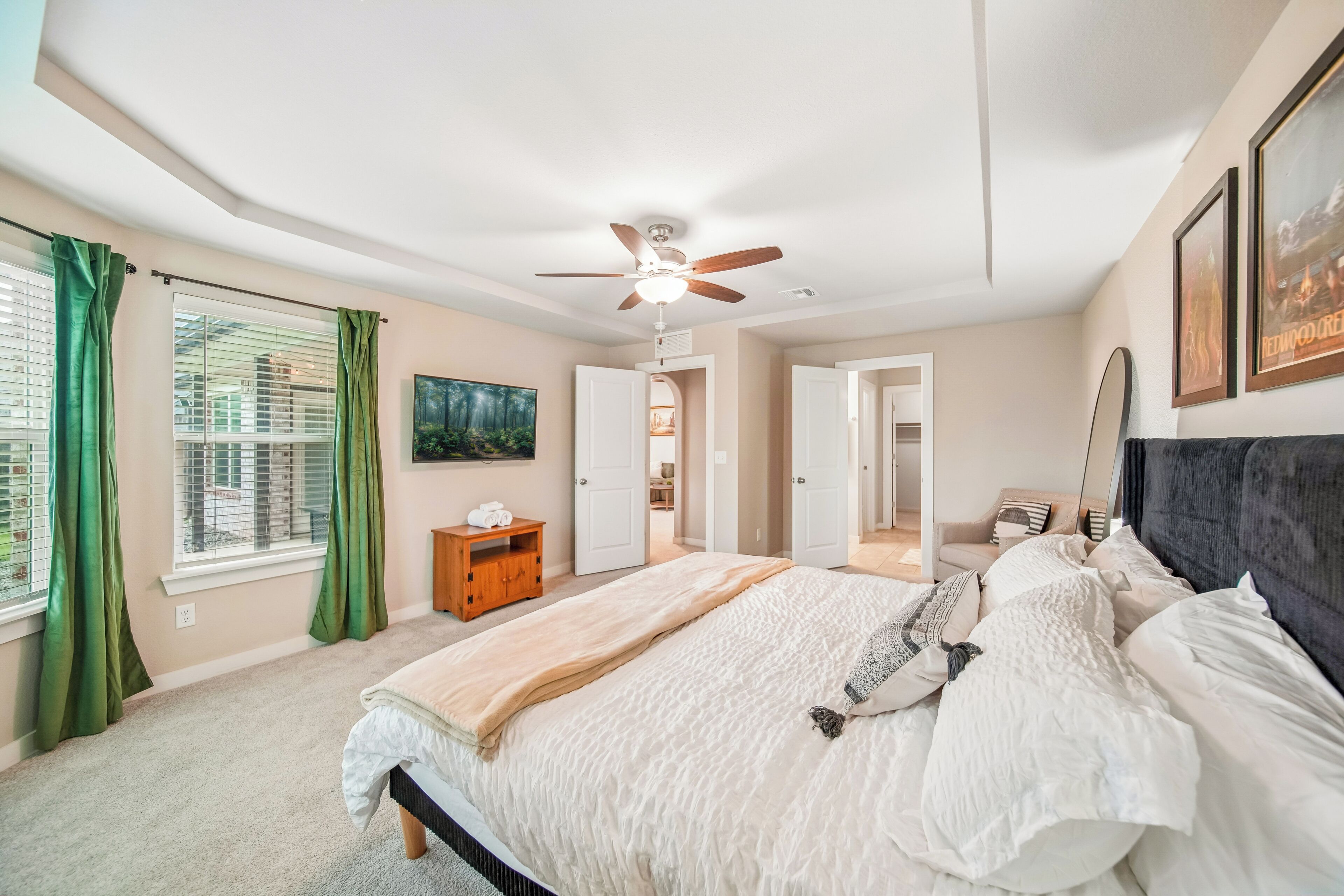Un dormitorio con cama, ventilador de techo, ventana con cortinas, una puerta y un cuadro en la pared.