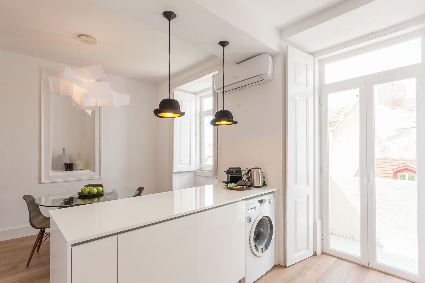 A modern kitchen with a central island, pendant lighting, a built-in washing machine, and large windows.