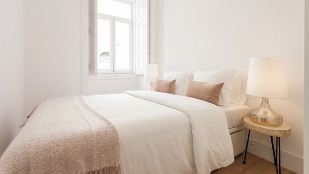 A minimalist bedroom with a large bed, a bedside table with a lamp, and a window providing natural light. - Your Lisbon Home Santa Apolónia (Lisbon)
