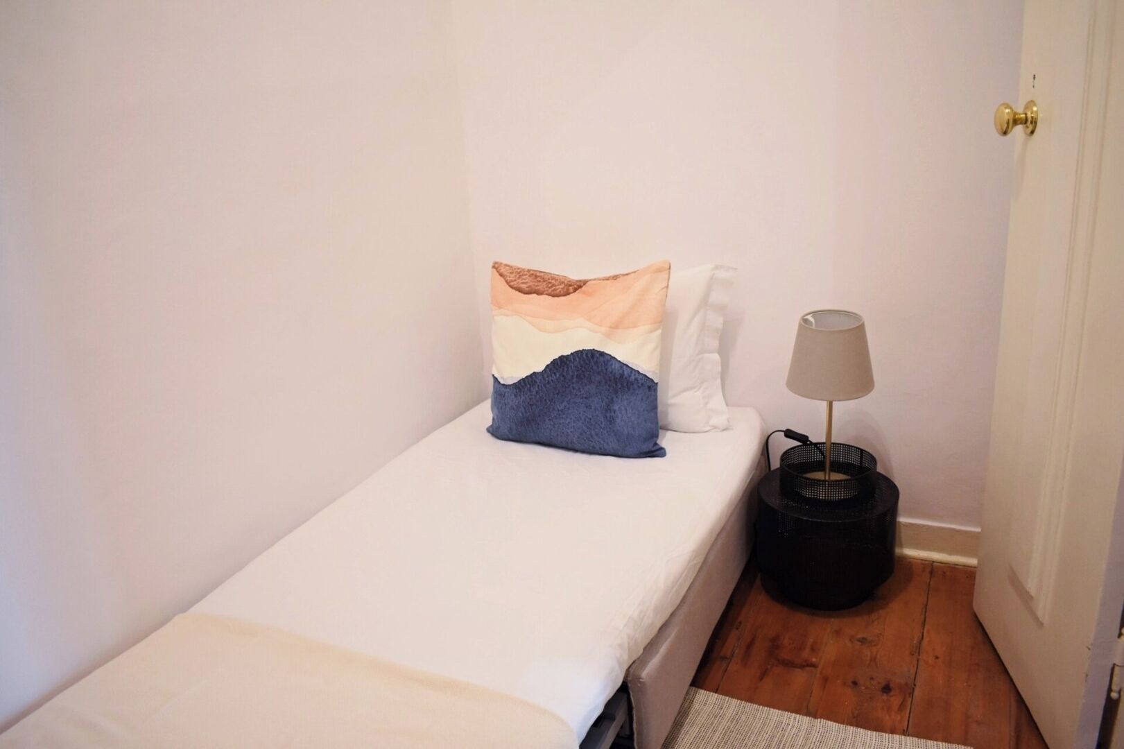 A small, single-bed room with a white bedspread, a blue pillow, and a lamp on a black side table.