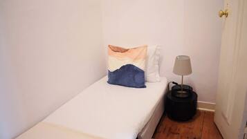 A small, single-bed room with a white bedspread, a blue pillow, and a lamp on a black side table.