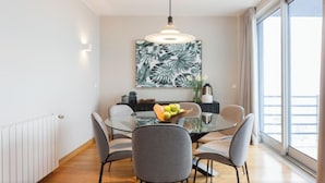 Et moderne spiseområde med et glasbord, grå stole, en skål med frugt og et stort maleri af et tropisk blad på væggen.