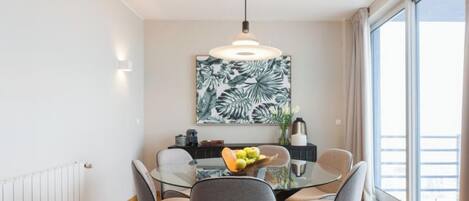 A modern dining area with a glass table, grey chairs, a fruit bowl, and a large tropical leaf painting on the wall.