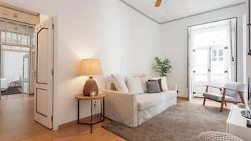A modern living room with a white sofa, a round wooden side table with a lamp, a potted plant, and a ceiling fan.