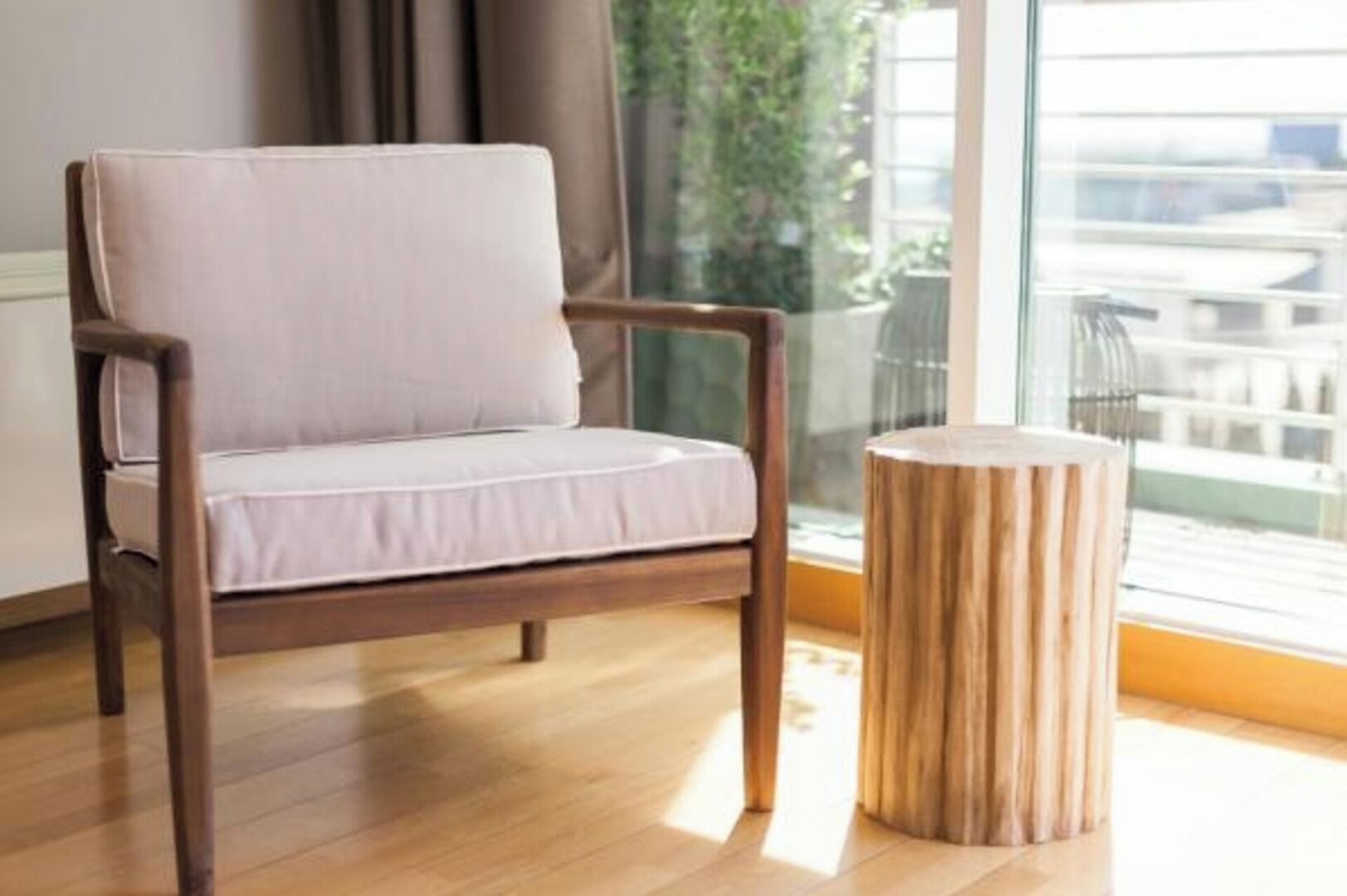 Une chaise à ossature en bois, dotée d’un coussin, est placée à côté d’un tabouret en bois dans une pièce avec une grande fenêtre.