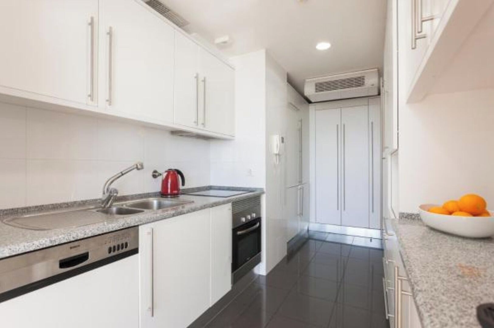 A modern kitchen with white cabinets, a stainless steel sink, a red kettle, and a bowl of oranges on the counter.