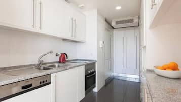 A modern kitchen with white cabinets, a stainless steel sink, a red kettle, and a bowl of oranges on the counter.