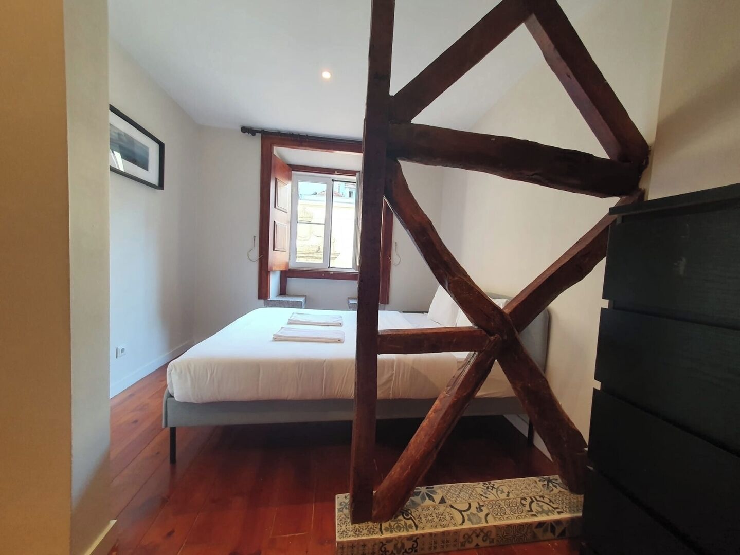 A bedroom with a wooden truss ceiling, a bed with white bedding, and a window providing natural light.