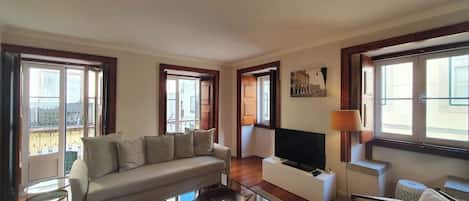 A living room with a sofa, a coffee table, a TV, and a lamp. There are windows with curtains and a painting on the wall.