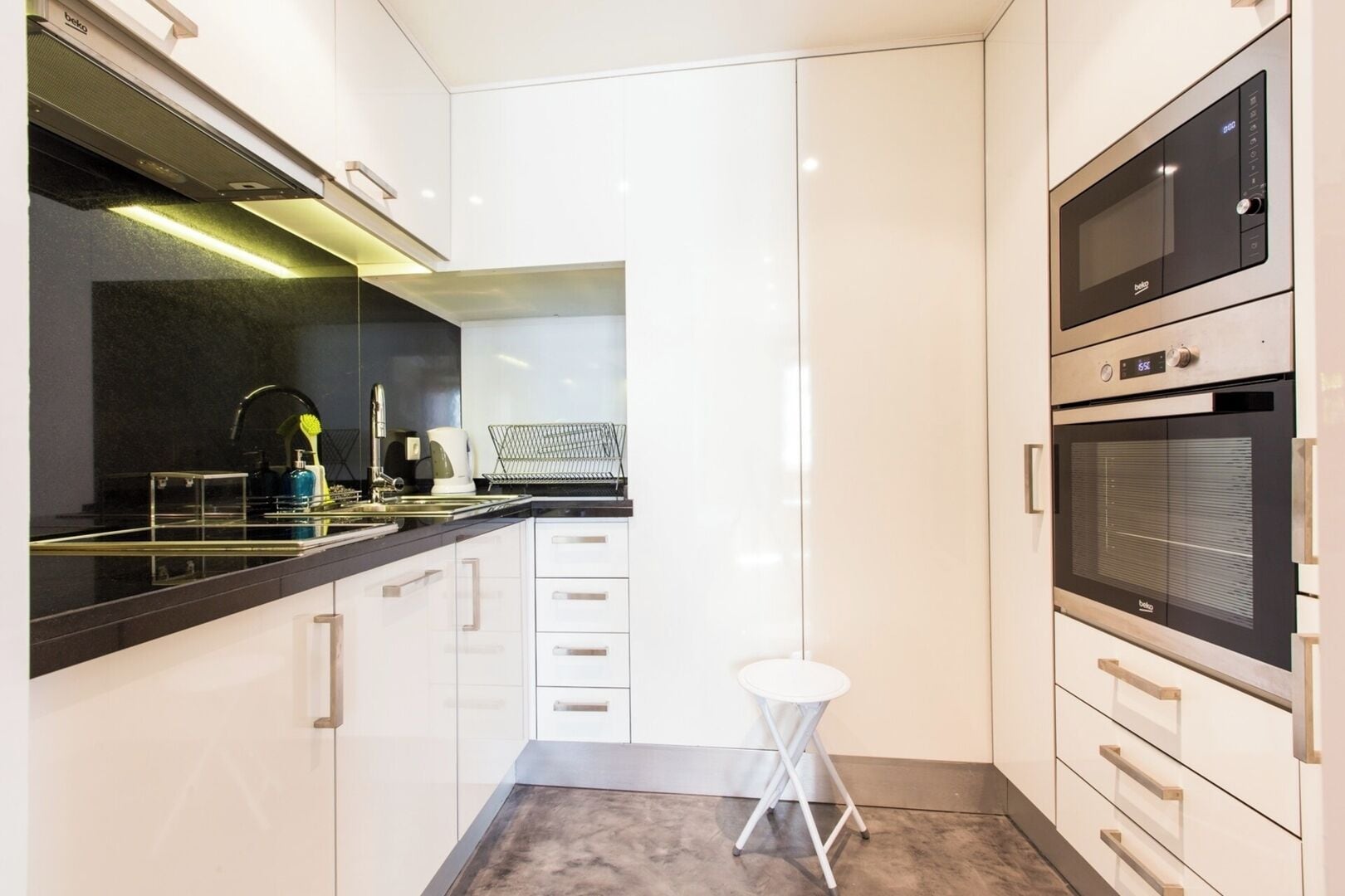 A modern kitchen with a black countertop, white cabinets, a stainless steel range hood, a built-in oven, and a small white stool.