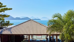 A beachside building with a tiled roof, outdoor seating, and a view of the sea and mountains. - VILLA NK SUITES (Ubatuba)