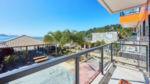 A balcony with a view of a beach, a covered area with tables, and a building with a mural. - VILLA NK SUITES (Ubatuba)