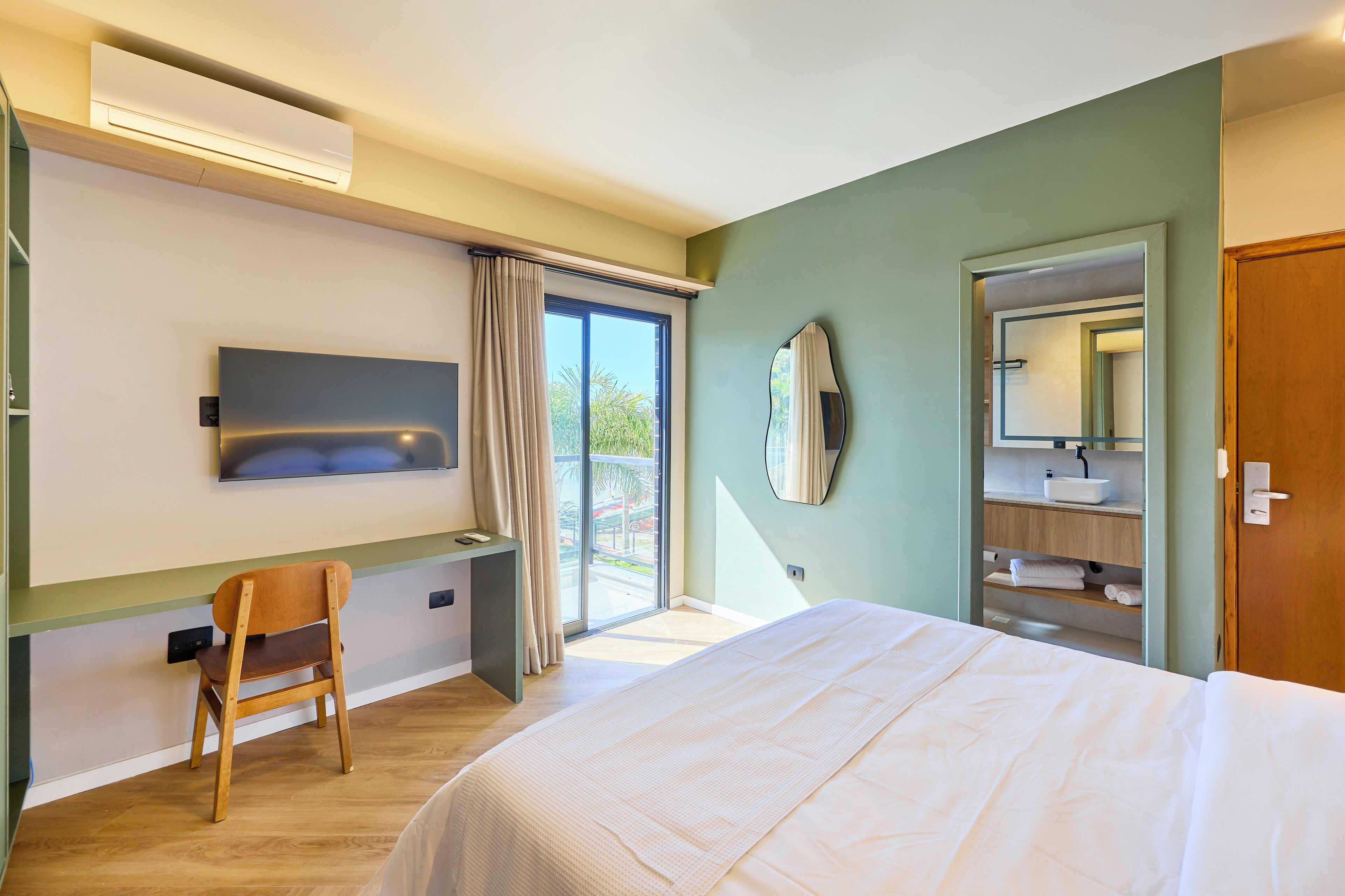Hotelkamer met een bed, bureau, stoel, groot raam met gordijnen, spiegel aan de muur en badkamer met wastafel en handdoekhouder.