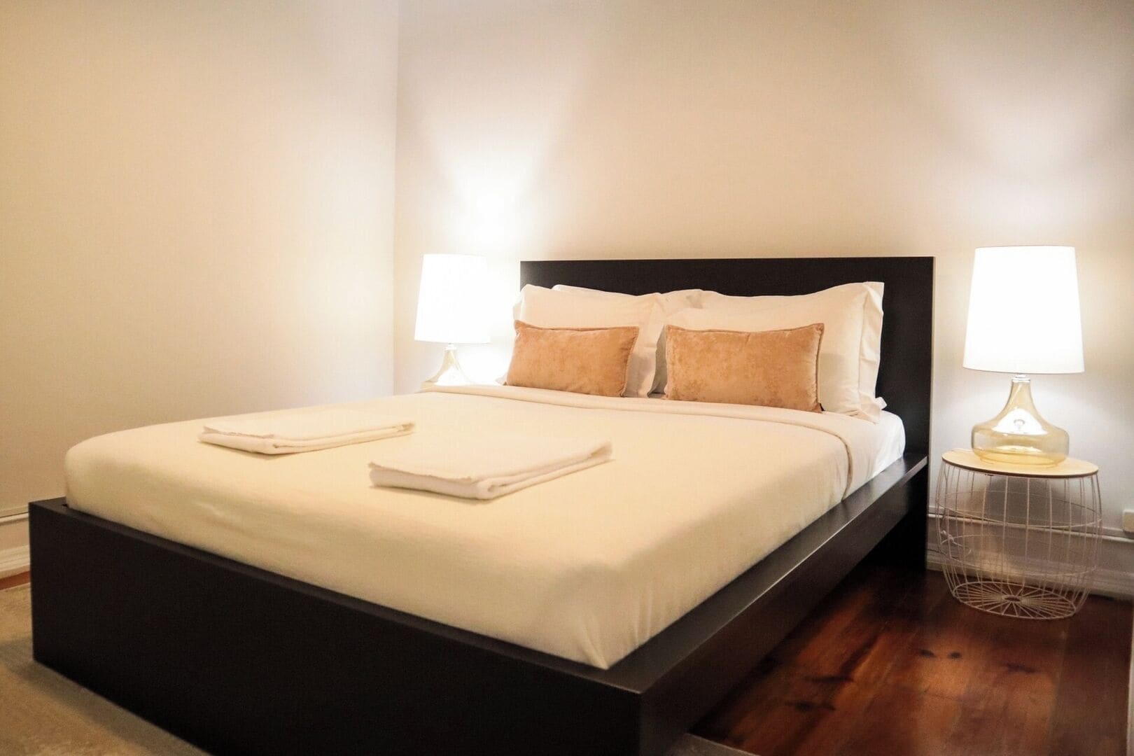 A neatly made bed with pillows and folded towels, a dark headboard, and two bedside lamps on a wooden floor.