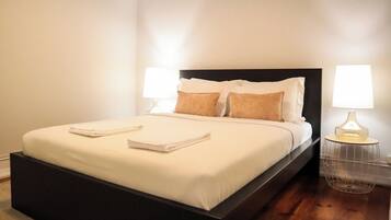 A neatly made bed with pillows and folded towels, a dark headboard, and two bedside lamps on a wooden floor.