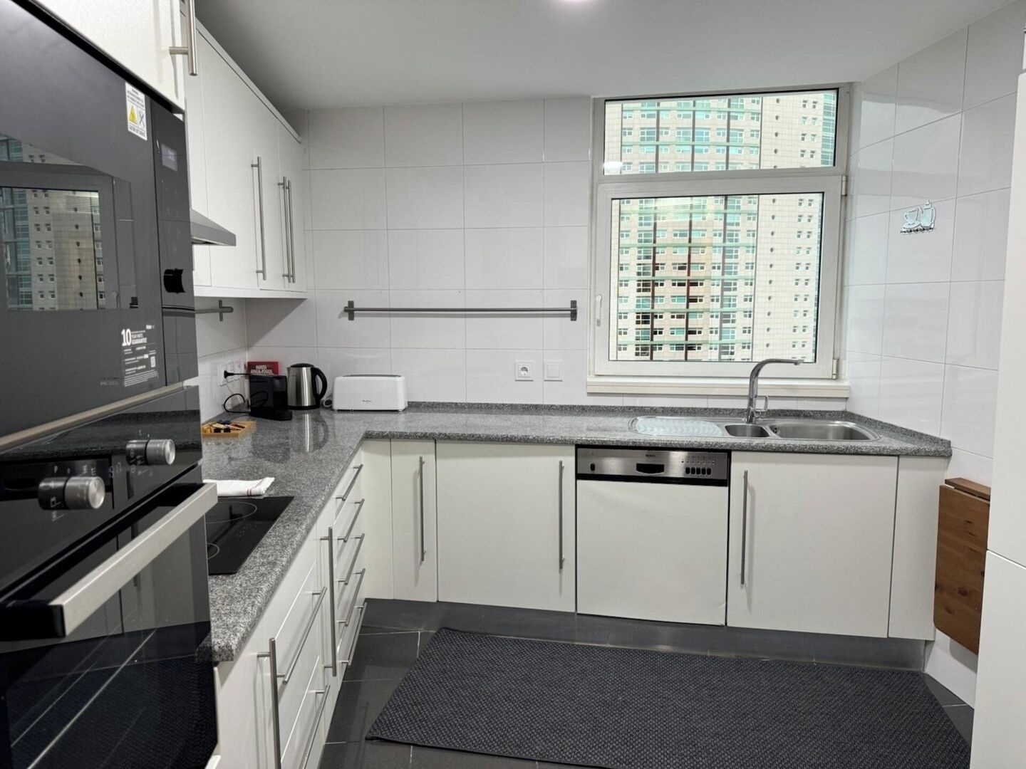 Cocina moderna con gabinetes blancos, refrigerador de acero inoxidable, lavavajillas integrado y ventana con vista a la ciudad.
