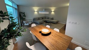 A modern living room with a wooden dining table, white chairs, a grey sectional sofa, and a large window providing a view of a bridge.