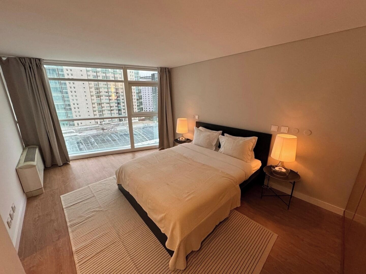 Habitación de hotel con una cama grande, dos lámparas de noche, vista a un paisaje urbano a través de un ventanal amplio y un radiador en la pared.