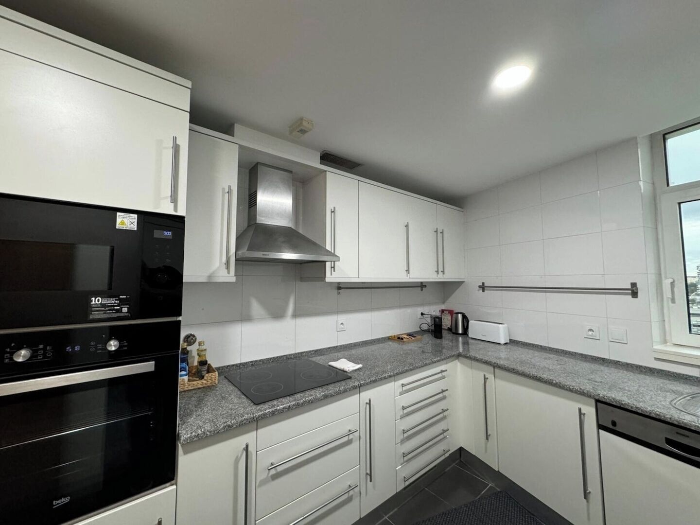 A modern kitchen with white cabinets, a stainless steel range hood, a built-in oven, and a window providing natural light.
